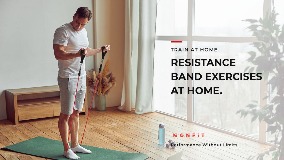 A man in a white tee stands on a green mat beside a floor to ceiling window and pulls against red MONFIT resistance tube bands anchored under his feet, on a timber floor.